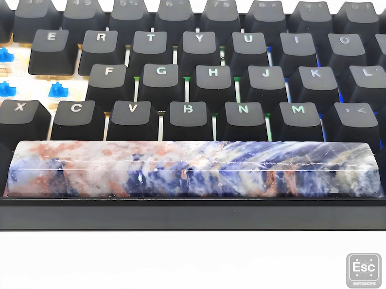 May include: A close-up of a mechanical keyboard with black keycaps. The spacebar is a custom design with a marbled pattern in shades of blue, white, and red. The keyboard has blue and green LED lights.