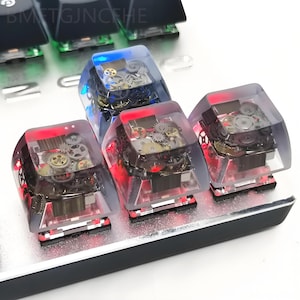 Puede incluir: Primer plano de cuatro teclas de teclado mecánico personalizadas. Cada tecla está encapsulada en resina transparente, mostrando intrincados engranajes de relojería e iluminación LED roja o azul. Las teclas se asientan sobre una base de teclado plateada.