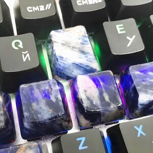 May include: Close-up of a mechanical keyboard with custom keycaps. The keycaps are made of a translucent material with a blue and white marbled pattern. The keyboard keys are black with white and blue lettering.