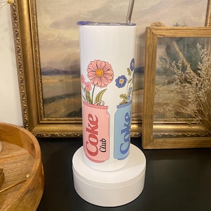 May include: A white tumbler with a floral design featuring pink and blue flowers. The tumbler has a silver straw and a lid. The design includes the words "Coke Club" on pink and blue cans.