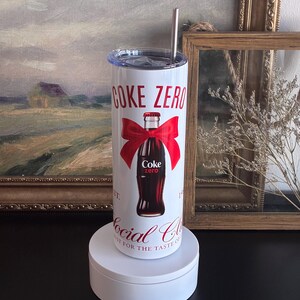 May include: A white stainless steel tumbler with a lid and a silver straw. The tumbler features the words "Coke Zero" in red, a red bow, and a Coke Zero bottle graphic. The words "Social Club" are also printed on the tumbler.
