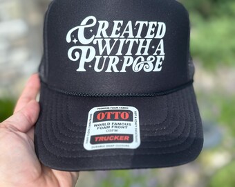 May include: Black trucker hat with the text "CREATED WITH A PURPOSE" in white script. The hat features a foam front and a mesh back. The brand label "OTTO" is visible.