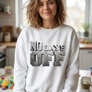 May include: A white sweatshirt with the text "NO DAYS OFF" in a metallic, textured font. The sweatshirt is long-sleeved and appears to be made of a soft material. The person wearing the sweatshirt is smiling.