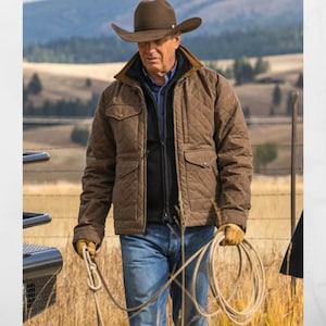 Op de afbeelding: Een bruine gewatteerde jas met een corduroy kraag, gedragen over een zwarte jas en een blauw overhemd. De persoon draagt een bruine cowboyhoed en een blauwe spijkerbroek en houdt een touw vast. De achtergrond toont een veld en een hek.