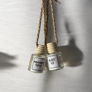 Può includere: Due piccoli flaconi in vetro trasparente per profumatori d'auto con tappi in legno chiaro e cordini intrecciati color beige. Ogni flacone presenta un'etichetta rettangolare bianca con bordo smerlato. Un'etichetta riporta Mahogany Fierce e l'altra Black Ice in nero.