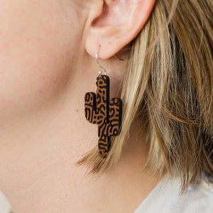 May include: A pair of brown wooden earrings shaped like a cactus with a black and brown patterned design. The earrings are hanging from a person's ear.