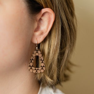 May include: A pair of wooden earrings with a geometric pattern. The earrings are brown with a black design and have a triangle shape with a cutout in the center.
