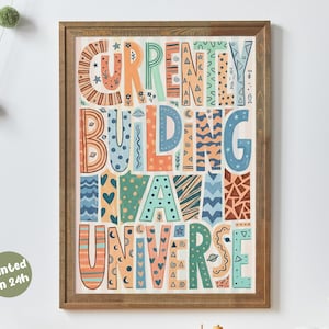 Op de afbeelding: Een ingelijste kunstafdruk met de tekst "Currently Building My Own Universe" in kleurrijke, patroonletters. De print heeft een houten lijst en een witte achtergrond. De zin is in verschillende kleuren, waaronder oranje, blauw en groen.
