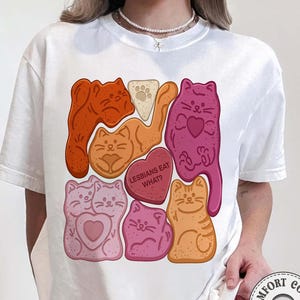 Puede incluir: Camiseta blanca con un gráfico de galletas con forma de gato en tonos naranja, rosa y morado. Una galleta tiene forma de corazón con el texto "LESBIANS EAT WHAT?". La camiseta tiene un corte holgado y está hecha de un material suave.