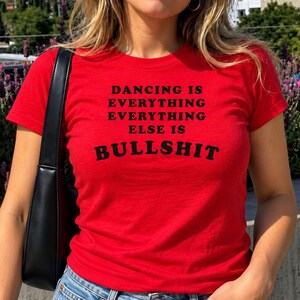 Könnte beinhalten: Ein leuchtend rotes Kurzarm-T-Shirt mit schwarzem Text, der DANCING IS EVERYTHING EVERYTHING ELSE IS BULLSHIT lautet. Das Shirt wird mit einer schwarzen Umhängetasche und einer blauen Jeans kombiniert, was einen lässigen Look mit Schriftzug ergibt.