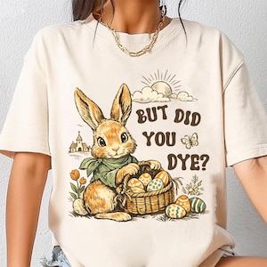 May include: A cream-coloured t-shirt with an Easter-themed graphic. It shows a cartoon rabbit with a basket of decorated eggs, the text "BUT DID YOU DYE?", a sun, clouds, and a church. The rabbit is wearing a green scarf.
