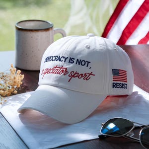 May include: White baseball cap with the text "DEMOCRACY IS NOT, a spectator sport" in red and black embroidery. Features an American flag patch with the word "RESIST". Includes a pair of sunglasses and a coffee mug.
