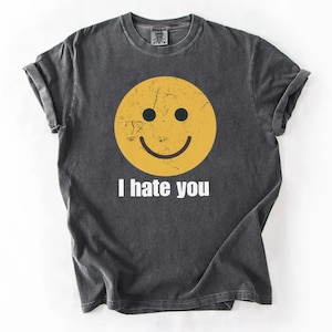 Può includere: T-shirt grigio scuro con una faccina sorridente gialla consumata e la scritta bianca "I hate you". La maglietta ha le maniche arrotolate e il girocollo.