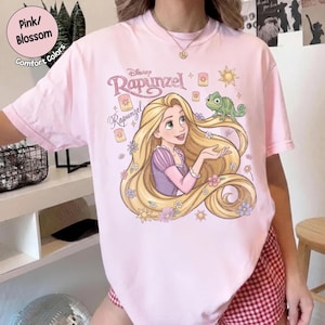 May include: A light pink t-shirt featuring a graphic print of the character Rapunzel with long blonde hair, a green chameleon, and floating lanterns. The text reads Disney Rapunzel. The shirt has short sleeves and a crew neckline, showcasing a colorful illustration.