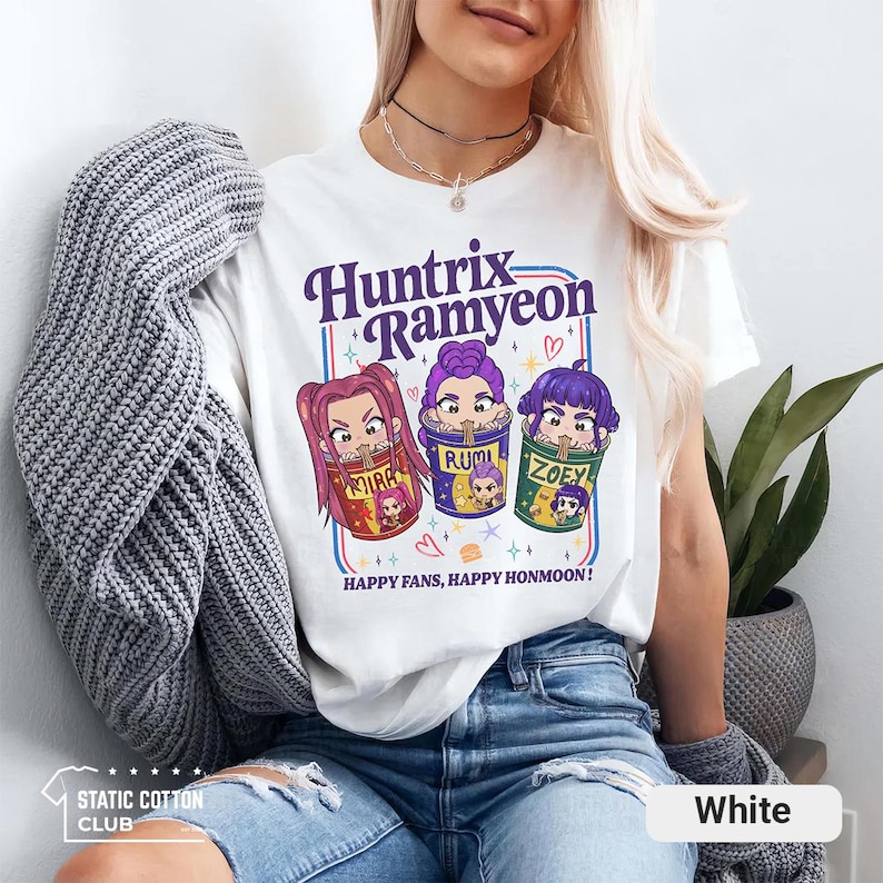 May include: White t-shirt featuring the text "Huntrix Ramyeon" in purple, with three cartoon characters in colorful cups. The text "HAPPY FANS, HAPPY HONMOON!" is below the graphic. The shirt is from Static Cotton Club.