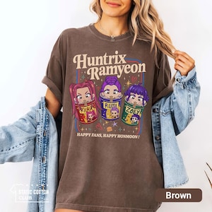 May include: Brown t-shirt with the text "Huntrix Ramyeon" and cartoon illustrations of three characters in ramen bowls. The shirt also has the text "HAPPY FANS, HAPPY HONMOON!" printed on it. The shirt is a relaxed fit.