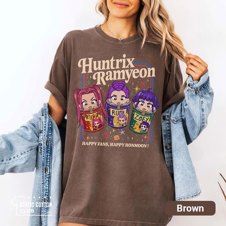 May include: Brown t-shirt with the text "Huntrix Ramyeon" and cartoon illustrations of three characters in ramen bowls. The shirt also has the text "HAPPY FANS, HAPPY HONMOON!" printed on it. The shirt is a relaxed fit.