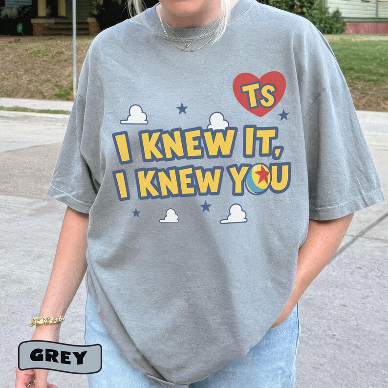 K&ouml;nnte beinhalten: Ein graues Kurzarm-T-Shirt mit einem Grafikdruck, der den Text "I KNEW IT, I KNEW YOU" in gelben Buchstaben mit blauen Umrandungen zeigt. Das Design enth&auml;lt ein rotes Herz mit "TS" darin, wei&szlig;e Wolken, blaue Sterne und einen bunten Ball.