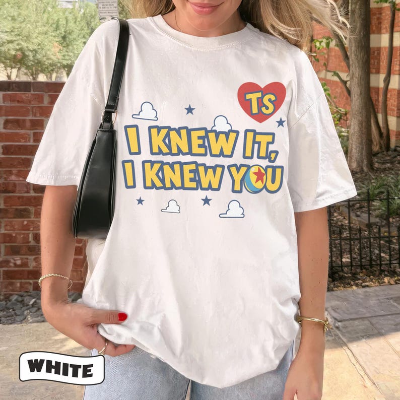 K&ouml;nnte beinhalten: Ein wei&szlig;es T-Shirt mit kurzen &Auml;rmeln, das ein rotes Herz mit den Buchstaben TS, den Text I KNEW IT, I KNEW YOU und eine Grafik eines Balls mit einem roten Stern zeigt, umgeben von blauen Wolken- und Sternsymbolen. Ein schwarzer Schultertaschenriemen ist sichtbar.