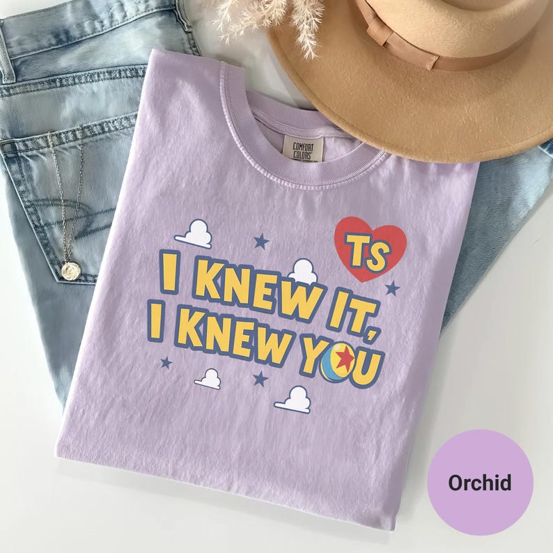 K&ouml;nnte beinhalten: Ein hellviolettes T-Shirt mit dem Text I KNEW IT, I KNEW YOU in Gelb mit blauen Umrandungen. Das Design enth&auml;lt ein rotes Herz mit den Buchstaben TS, kleine wei&szlig;e Wolken, blaue Sterne und eine bunte Kugelgrafik mit einem roten Stern.