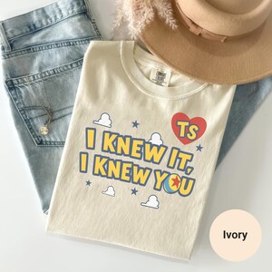 K&ouml;nnte beinhalten: Ein elfenbeinfarbenes T-Shirt mit dem blauen Schriftzug I KNEW IT, I KNEW YOU, einem roten Herz mit den Buchstaben TS, kleinen blauen Sternen und wei&szlig;en Wolkenumrissen. Das T-Shirt liegt neben einer hellblauen Jeans und einem beigen Filzhut.