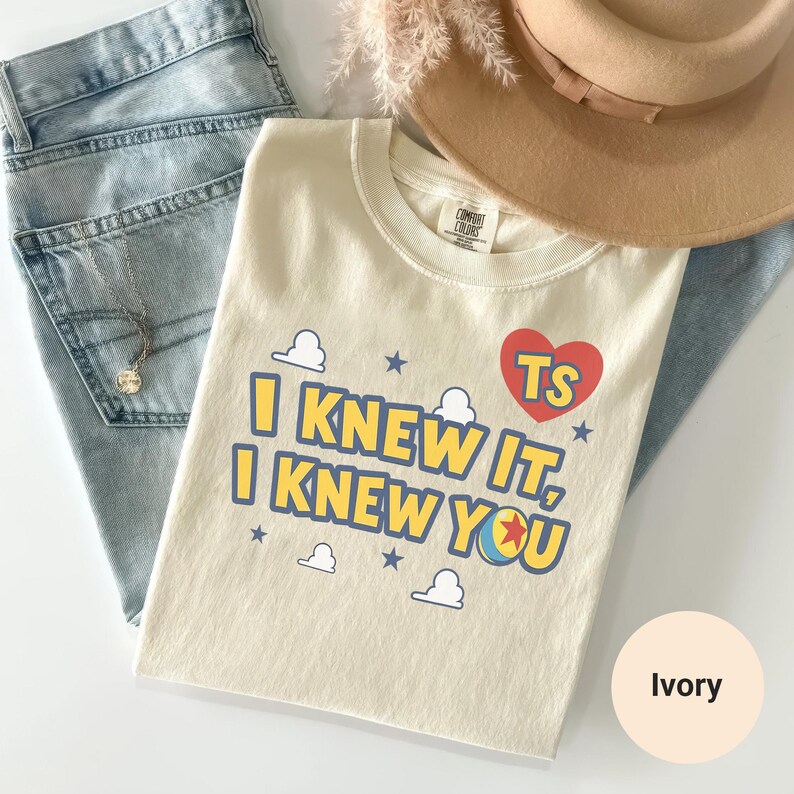 K&ouml;nnte beinhalten: Ein elfenbeinfarbenes T-Shirt mit dem blauen Schriftzug I KNEW IT, I KNEW YOU, einem roten Herz mit den Buchstaben TS, kleinen blauen Sternen und wei&szlig;en Wolkenumrissen. Das T-Shirt liegt neben einer hellblauen Jeans und einem beigen Filzhut.