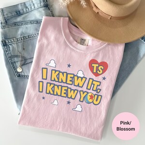 K&ouml;nnte beinhalten: Ein hellrosa T-Shirt mit dem Text I KNEW IT, I KNEW YOU in Gelb mit blauen Umrandungen, einem roten Herz mit TS, einem bunten Ball sowie kleinen wei&szlig;en Wolken- und blauen Sternmotiven. Das T-Shirt liegt neben einer hellblauen Jeans und einem beigen Hut.