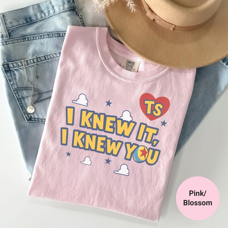 K&ouml;nnte beinhalten: Ein hellrosa T-Shirt mit dem Text I KNEW IT, I KNEW YOU in Gelb mit blauen Umrandungen, einem roten Herz mit TS, einem bunten Ball sowie kleinen wei&szlig;en Wolken- und blauen Sternmotiven. Das T-Shirt liegt neben einer hellblauen Jeans und einem beigen Hut.