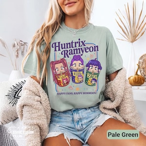 May include: Pale green t-shirt featuring the text "Huntrix Ramyeon" above three cartoon characters in ramen bowls. The shirt also has the text "HAPPY FANS, HAPPY HONMOON!". The shirt is made of a soft material.