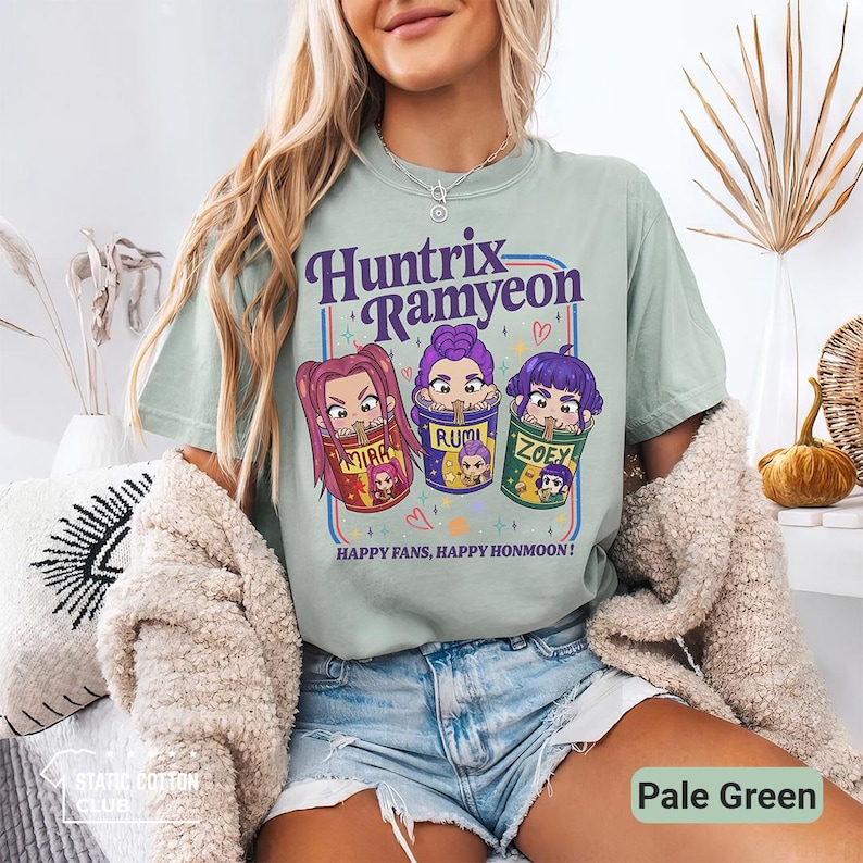 May include: Pale green t-shirt featuring the text "Huntrix Ramyeon" above three cartoon characters in ramen bowls. The shirt also has the text "HAPPY FANS, HAPPY HONMOON!". The shirt is made of a soft material.