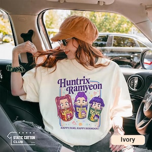 May include: Ivory-colored sweatshirt with the text "Huntrix Ramyeon" and cartoon illustrations of three characters in cups. The text "HAPPY FANS, HAPPY HONMOON!" is below the illustrations. The Static Cotton Club logo is in the lower left.