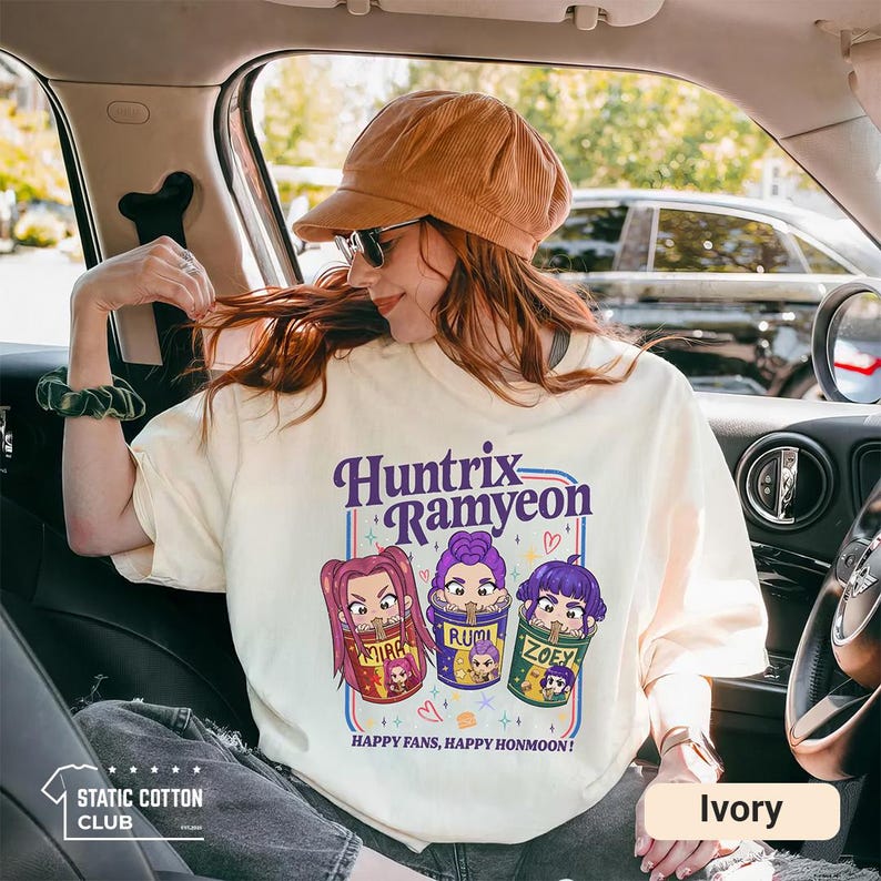May include: Ivory-colored sweatshirt with the text "Huntrix Ramyeon" and cartoon illustrations of three characters in cups. The text "HAPPY FANS, HAPPY HONMOON!" is below the illustrations. The Static Cotton Club logo is in the lower left.
