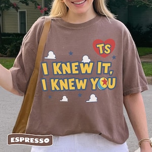 K&ouml;nnte beinhalten: Ein braunes Kurzarm-T-Shirt mit einem roten Herz mit den Buchstaben TS, dem Text I KNEW IT, I KNEW YOU und einer Grafik eines Balls mit einem roten Stern. Das Design enth&auml;lt kleine wei&szlig;e Wolken und blaue Sterne auf der Vorderseite.