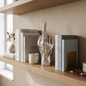 May include: A silver-coloured hand sculpture in a middle finger gesture positioned between two stacks of books. The books include titles such as Kinfolk, Atelier, and Less. A small white candle with gold accents sits next to the sculpture.