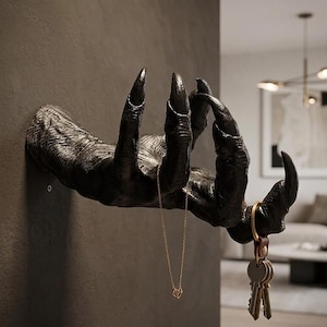 Könnte beinhalten: Ein schwarzer, klauenartiger Wandhaken, der einer Monsterhand ähnelt, ist an einer dunkelgrauen Wand befestigt. Eine goldene Halskette hängt an den Fingern, und ein Schlüsselbund ist am Handgelenk befestigt. Ein einzigartiger Dekorationsartikel.