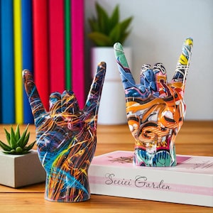 Pode incluir: Duas esculturas decorativas de mãos no gesto de rock. Uma é pintada com uma mistura vibrante de linhas azuis, laranja e amarelas. A outra apresenta um design em estilo grafite com várias cores. As esculturas estão sobre uma superfície de madeira.