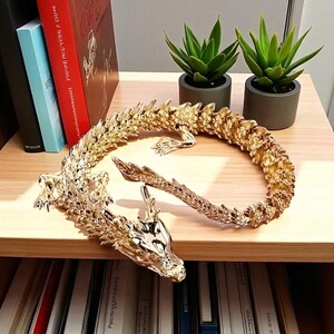 May include: A golden dragon figurine with intricate detailing, curled on a wooden shelf. The dragon's head is visible, along with its tail and body. Two small green potted plants are in the background, along with books.