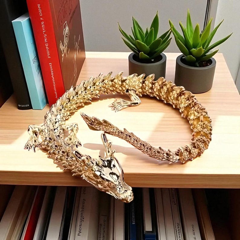 May include: A golden dragon figurine with intricate detailing, curled on a wooden shelf. The dragon's head is visible, along with its tail and body. Two small green potted plants are in the background, along with books.