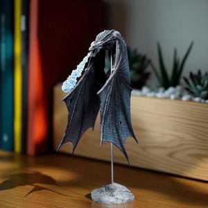 May include: A detailed dragon figurine with outstretched black wings and a blue flame effect emanating from its mouth. The dragon is mounted on a metal stand and a stone-like base. The figurine is displayed on a wooden surface.