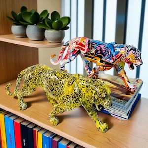 Peut inclure: Deux sculptures de léopards décoratives. L'une est jaune avec des taches noires, l'autre est peinte avec un design graffiti coloré. Elles sont posées sur une étagère en bois avec des livres et des plantes en pot.