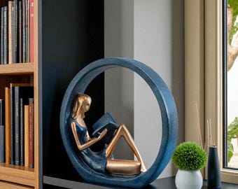 Könnte beinhalten: Dekorative Skulptur einer lesenden Frau in einem blauen, kreisförmigen Rahmen. Die Figur sitzt, mit goldfarbenen Beinen und einem blauen Kleid. Die Skulptur steht auf einem schwarzen Regal, in der Nähe eines Fensters und eines Bücherregals.