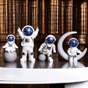 Puede incluir: Cuatro figuras de astronautas blancos con cascos azules estrellados. Una está sentada en una luna gris, otra está de pie con los brazos levantados, una tercera está arrodillada con una pequeña luna y la última está sentada en una luna creciente blanca. Las figuras se exhiben sobre una superficie blanca.