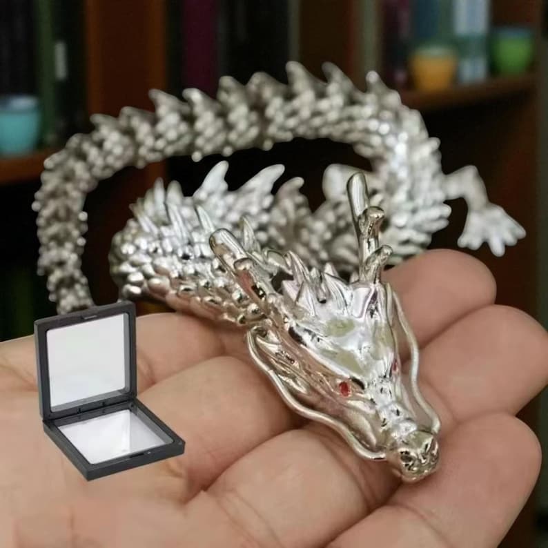May include: A detailed silver dragon figurine with red eyes and intricate scales. The dragon is partially coiled, with its head in the foreground. A small, open black display case is in the lower left corner. The dragon is a fantasy-themed collectible.