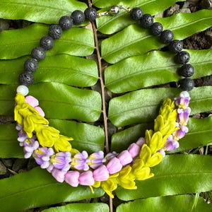 May include: A necklace with black lava rock beads and a floral design. The floral section has yellow and pink petals with purple accents. The necklace is set against a backdrop of green leaves.