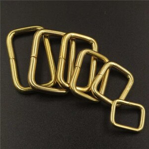 May include: A collection of five brass-colored, square-shaped metal rings. The rings are arranged in a staggered pattern, with each ring slightly smaller than the last. The metal has a shiny, reflective surface.