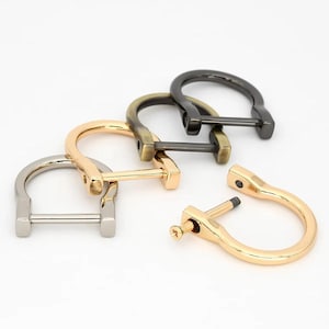 Op de afbeelding: Een assortiment metalen D-ring beugels in verschillende afwerkingen: zilver, goud, antiek messing en zwart. Eén beugel is gedemonteerd, met de schroef zichtbaar. Deze hardware componenten zijn waarschijnlijk voor leerbewerking of tassen maken.