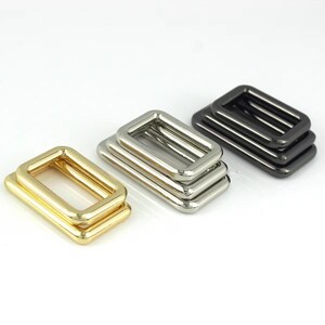 May include: Three sets of rectangular metal rings in gold, silver, and black. Each set comprises three stacked rings, displaying different finishes. These hardware components are likely used in bag making or other crafting projects.