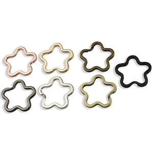 Può includere: Una collezione di portachiavi in metallo a forma di stella in vari colori, tra cui oro rosa, oro, bronzo, nero e argento. I portachiavi sono disposti su uno sfondo bianco.