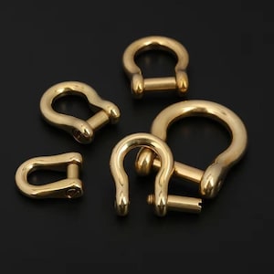 May include: A collection of gold-colored D-ring shackles in various sizes. The shackles are made of a shiny, metallic material and are arranged on a dark background. The D-ring shape is clearly visible, with a pin closure.