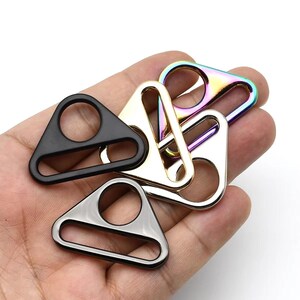 May include: A collection of metal triangle rings in various finishes: black, silver, gold, and iridescent rainbow. Each ring features a circular opening within the triangle shape, suitable for crafting or hardware applications.
